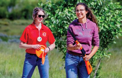 A female Game and Fish employee in a red shirt instructs a female hunter education student carrying an orange inert firearm outdoors.